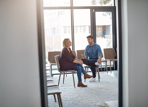 Two people sit in chairs on a rug in a modern room with large windows, engaged in a peaceful conversation. The room, designed for discussions of law and justice, has empty chairs lined up along the wall.