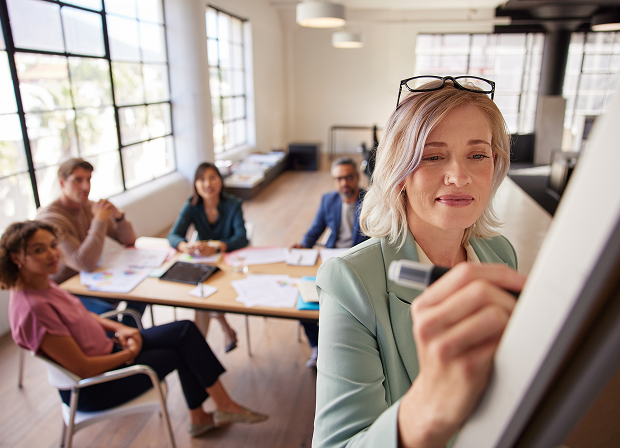A woman writes on a whiteboard about justice during a business meeting as four colleagues sit at a table in a modern office with large windows.