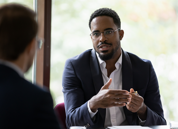 A man wearing glasses and a suit discusses matters of law with another person during a meeting, gesturing with his hand. The setting appears to be an office with a window in the background.
