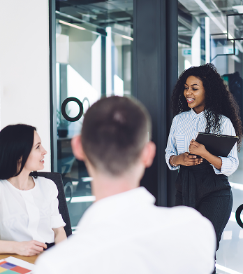 A woman holding a folder stands and smiles while speaking to two seated colleagues in a modern office meeting room. A woman holding a folder stands and smiles while speaking to two seated colleagues in a modern office meeting room.